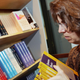 A woman holds a stack of yellow textbooks next to a bookshelf with a space laid out next to quantum mechanics.
